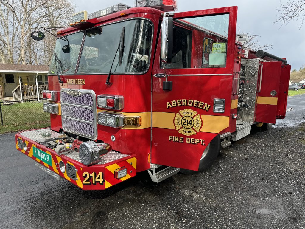 A red Aberdeen Fire Department fire truck with the number 214 is parked on a wet road, its door open. Trees, a house, and a fence are visible in the background.