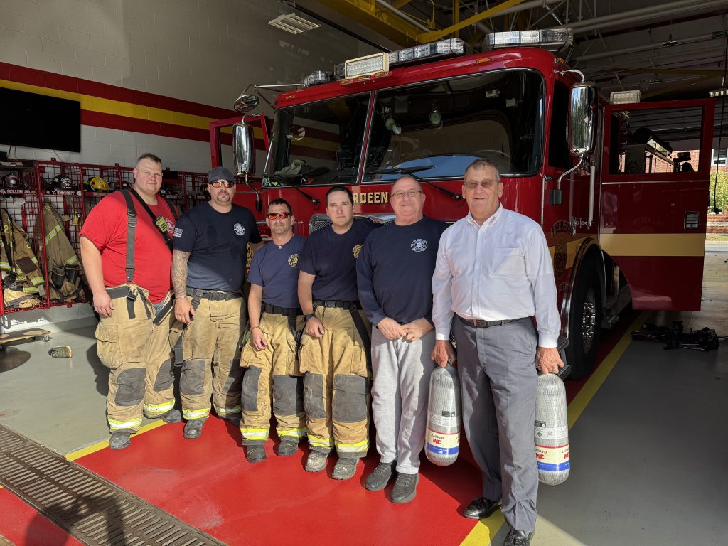 Six men, five in firefighting gear and one in business attire, stand in front of a red fire truck inside a firehouse. Two of the men are holding air tanks. Equipment and gear are visible in the background.