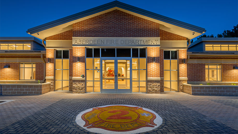 The Aberdeen Fire Department building at dusk, well-lit with large glass entrance doors, brick walls, and a fire department emblem marked “2” on the pavement in front.