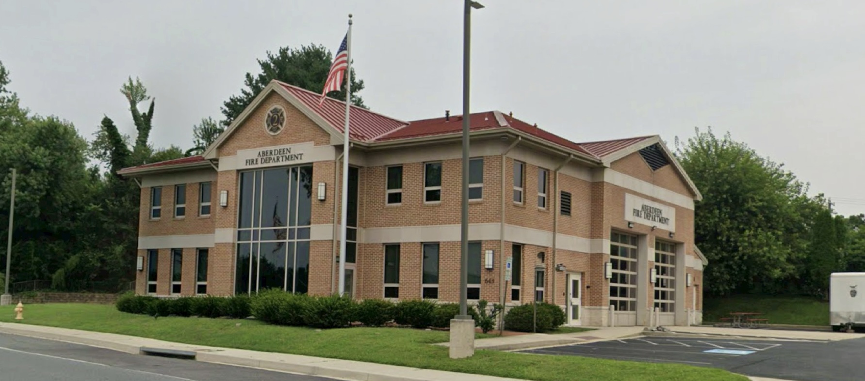 A two-story brick fire station with large windows and two garage doors, an American flag flying in front, and green trees surrounding the building.