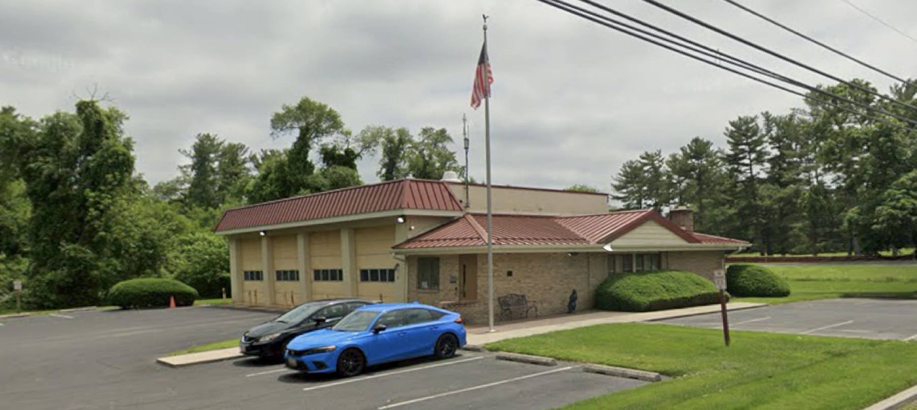 A one-story building with a red roof, flagpole, and three garage bays sits by a parking lot with two cars, surrounded by trees and a grassy area under a cloudy sky.