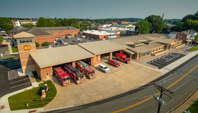 Aerial view of a fire station with several fire trucks parked inside and outside the garage bays, surrounded by parking lots and nearby buildings on a sunny day.
