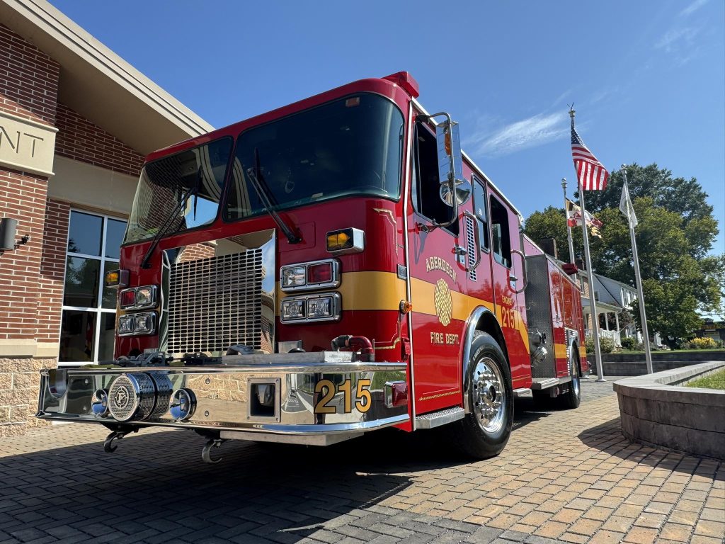 A red fire truck labeled “215” is parked outside a brick fire station on a sunny day, with American flags and trees visible in the background.