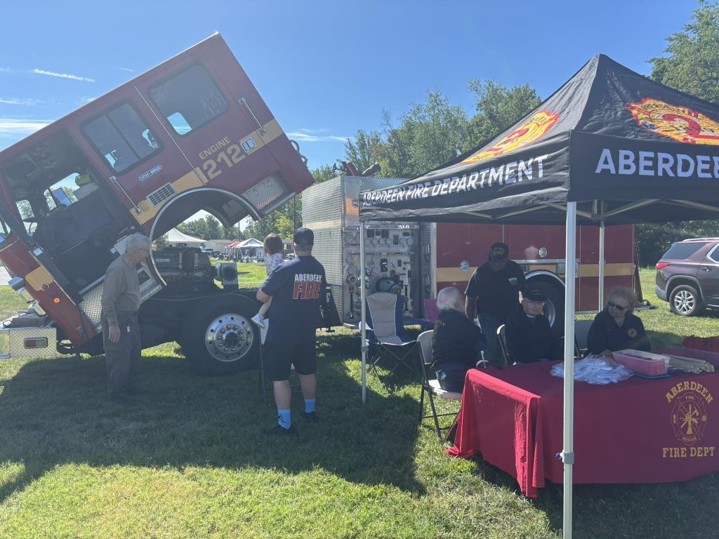 Aberdeen Fire Department members sit under a black tent with equipment on display next to a fire truck with its cab lifted. People visit the booth on a sunny day with trees in the background.