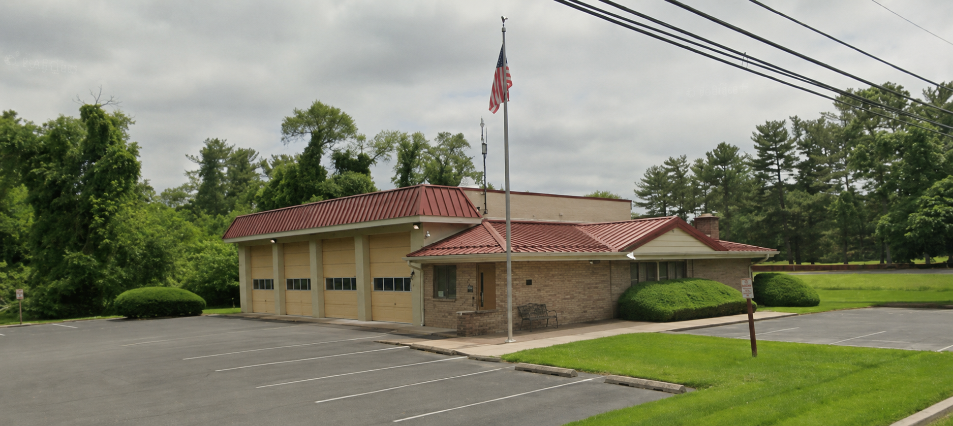 A small brick fire station with a red roof, three garage doors, and an American flag out front, surrounded by greenery and an empty parking lot.