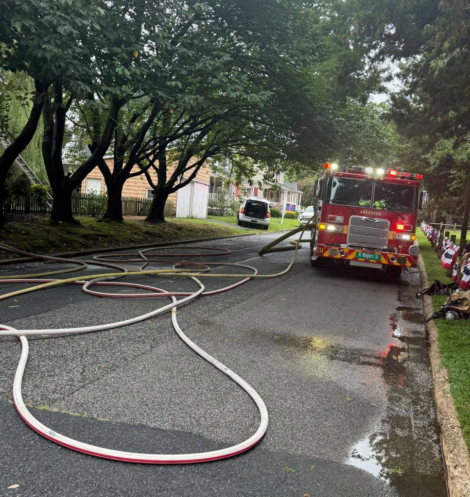 A fire truck with lights on is parked on a residential street. Multiple fire hoses are spread across the wet road, and a white vehicle is parked ahead. Trees and houses line both sides of the street.