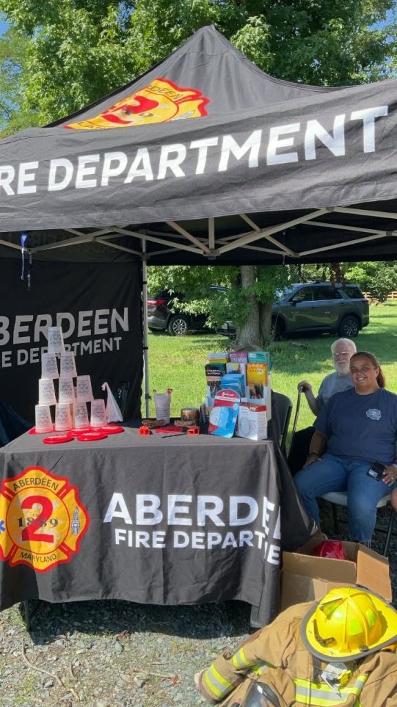 Aberdeen Fire Department tent with informational materials, water bottles, and helmets on display. Two people are sitting under the tent, smiling. Fire gear is on the ground in front. Trees and parked cars are in the background.